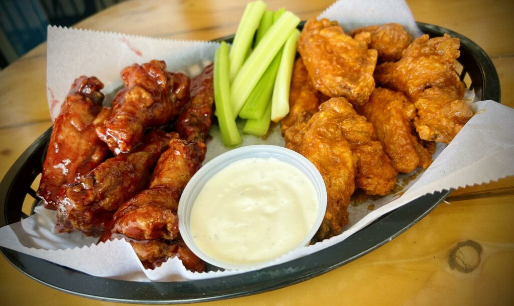 A basket of chicken wings, half covered in a dark sauce and half in a lighter orange sauce, served with celery sticks and a cup of ranch dressing on a parchment-lined tray.