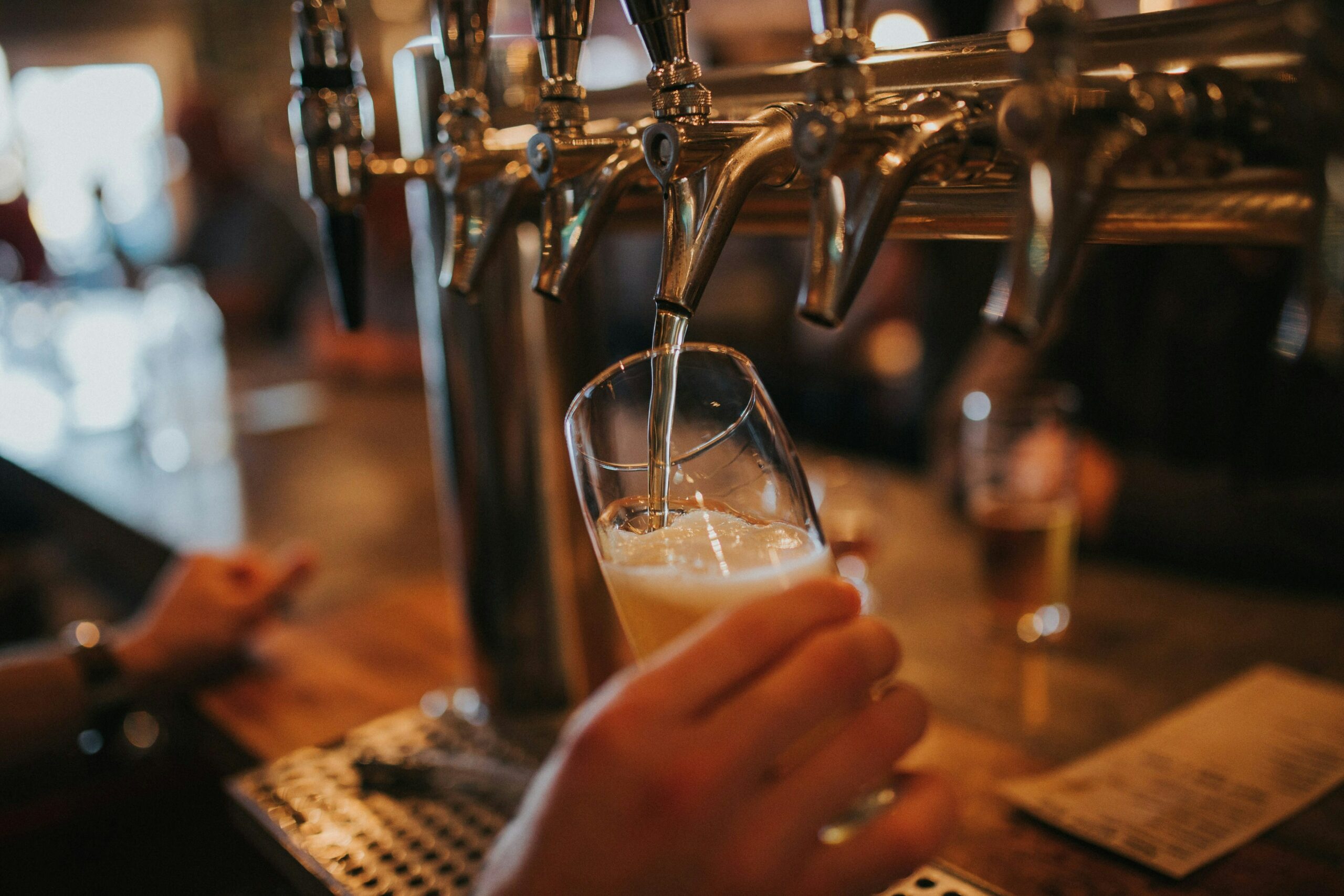 A hand holds a glass at an angle under a row of beer taps, filling it with draft beer in a dimly lit bar, while a menu and another glass are visible on the counter.