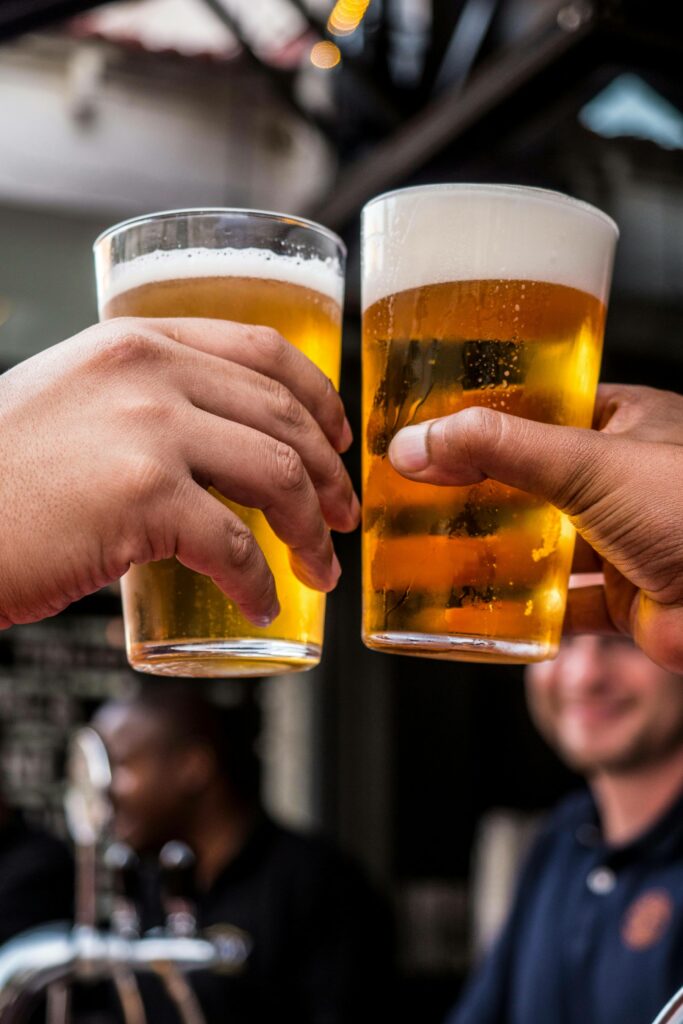 Two people clinking glasses of beer together in a celebratory toast, with a blurred background showing other people and a bar setting.