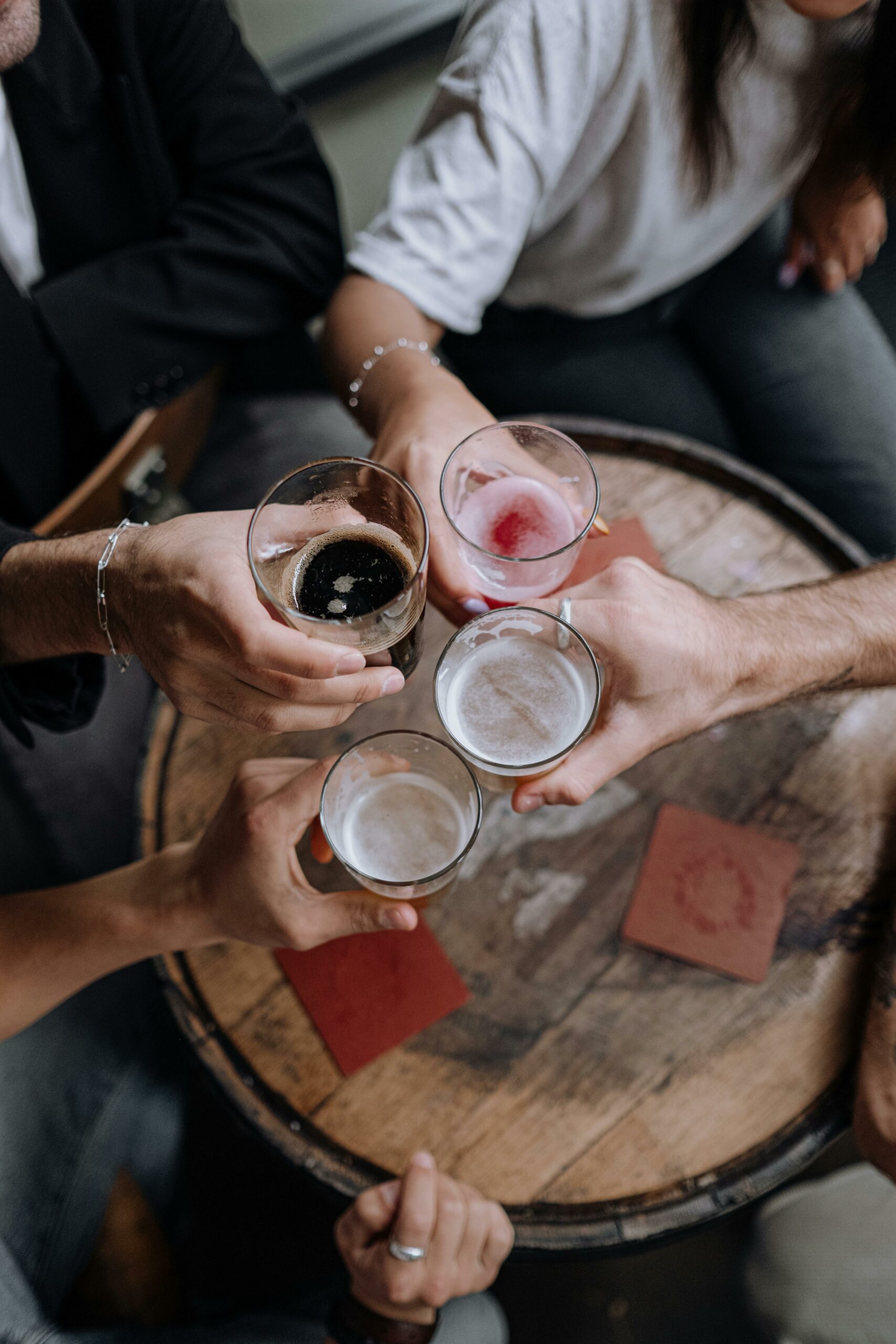 Four people clinking glasses of beer and cocktails over a round wooden table with red drink coasters, celebrating together from a top-down perspective.