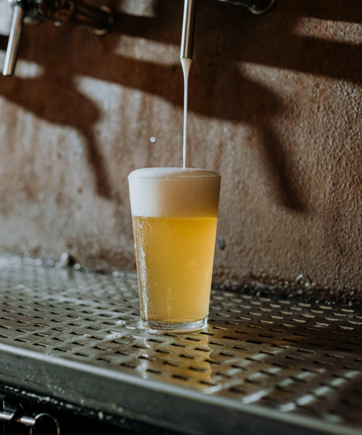 A glass of beer is being filled from a tap, creating a thick layer of foam on top. The glass sits on a metal drip tray beneath several beer taps, with a brown textured wall in the background.