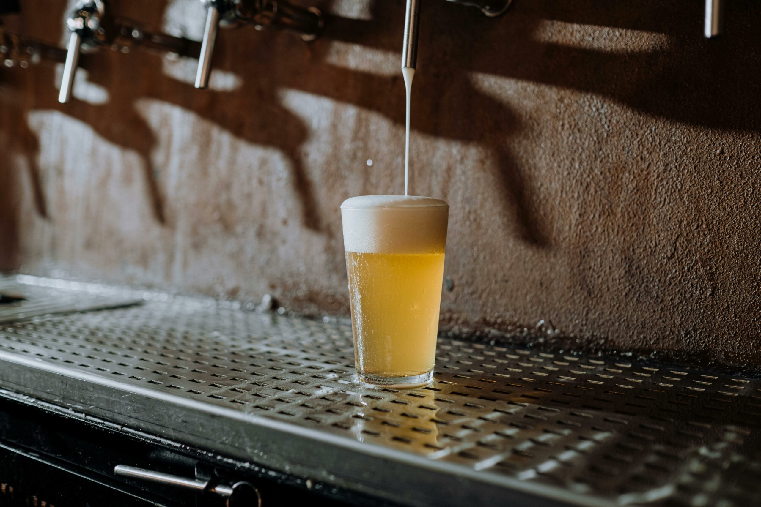 A glass of light beer being filled from a tap, with a thick foamy head forming on top, placed on a metal drip tray beneath beer taps in a bar setting.
