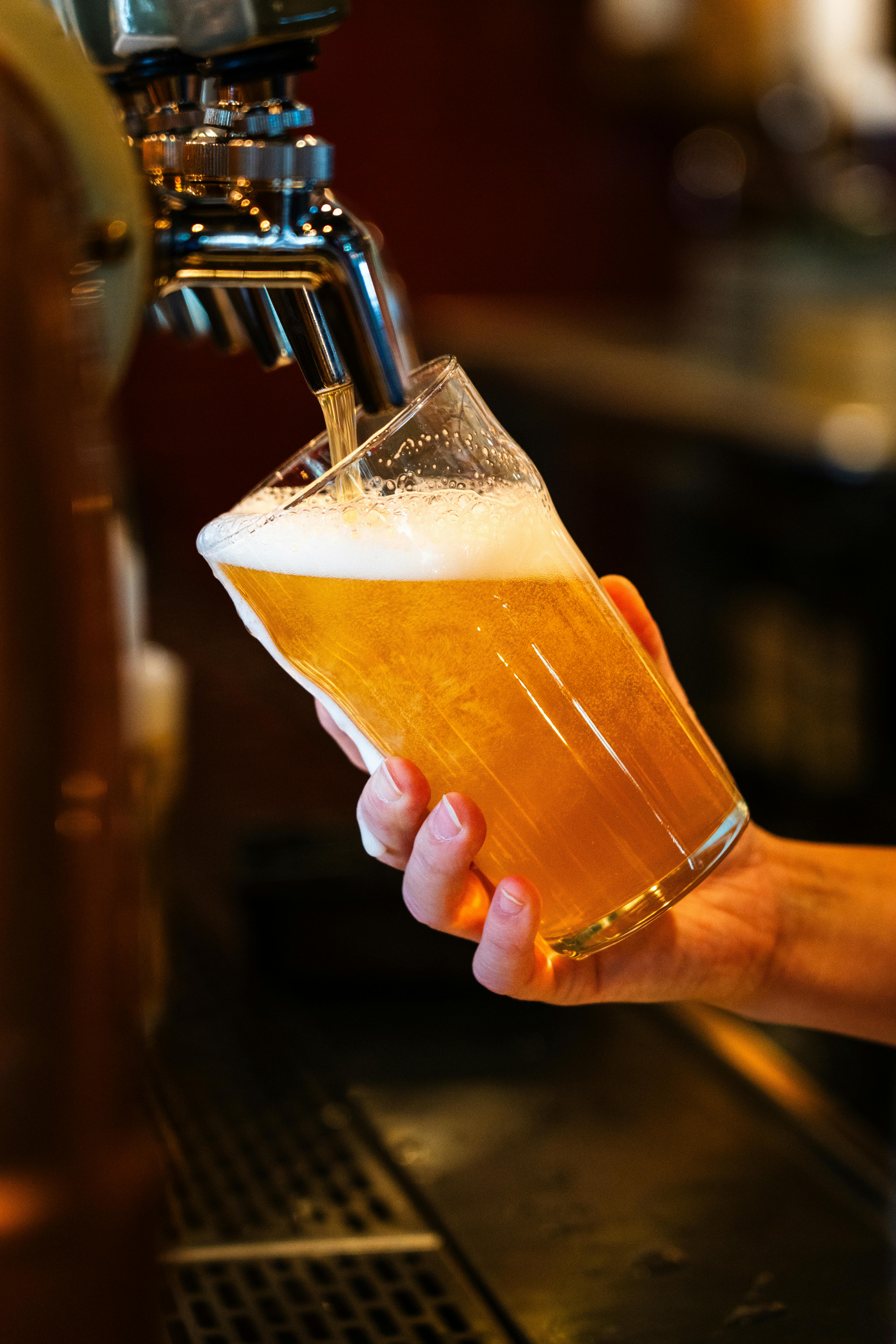 A hand holds a tilted glass as golden beer with a frothy head is poured from a tap at a bar.