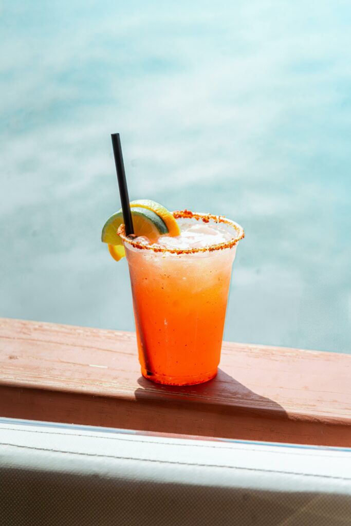 A bright orange cocktail in a plastic cup with a black straw, a lime wedge, and a chili-salt rim sits on a wooden ledge, with a blurred blue background suggesting water or a pool.