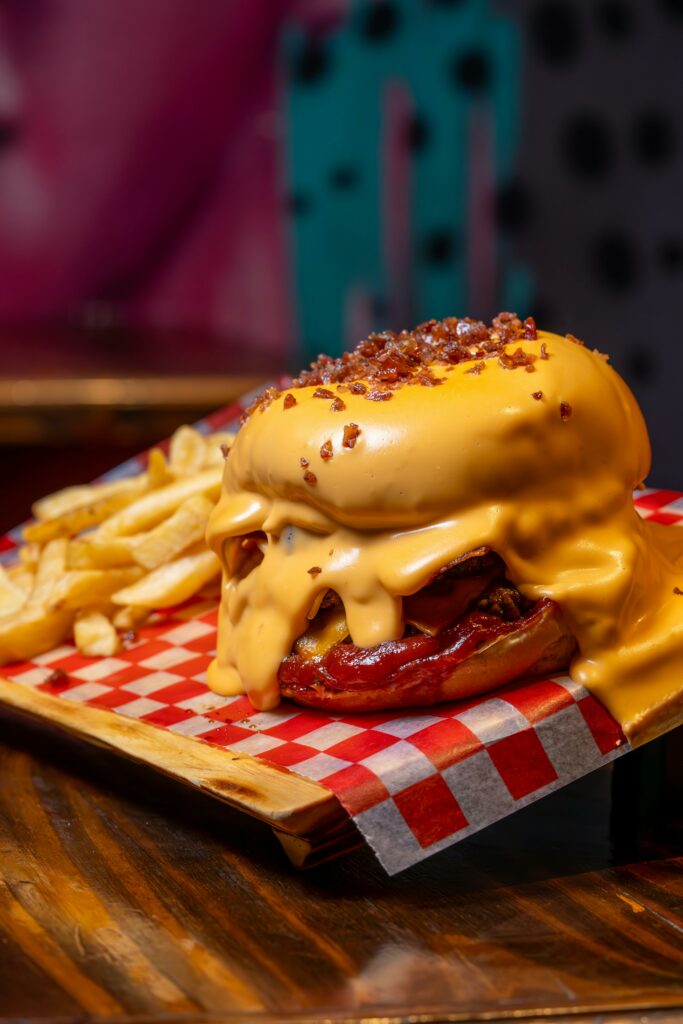 A cheeseburger covered in melted cheese sauce and bacon bits sits on a tray with French fries, atop red-and-white checkered paper. The cheese drips over the burger, nearly covering the whole sandwich.
