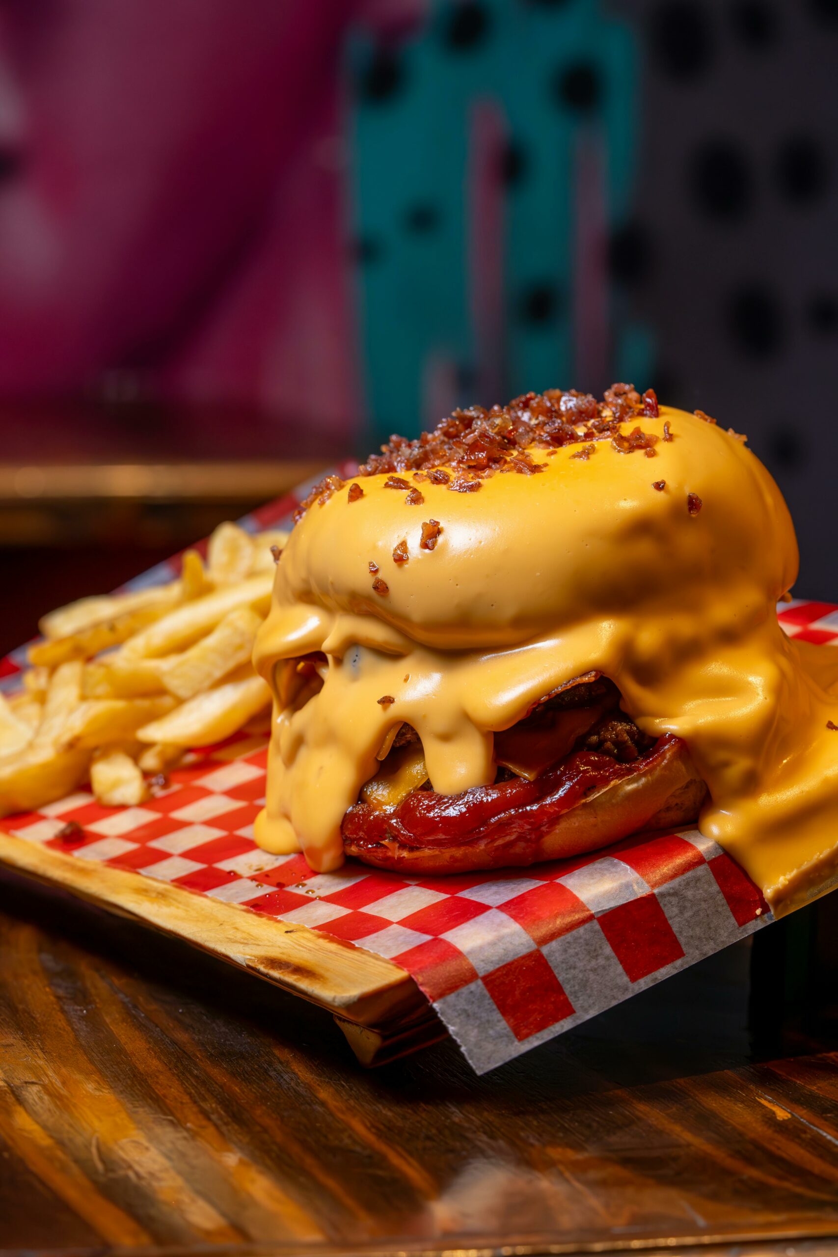 A cheeseburger covered in melted cheese sauce and bacon bits sits on a tray with French fries, atop red-and-white checkered paper. The cheese drips over the burger, nearly covering the whole sandwich.