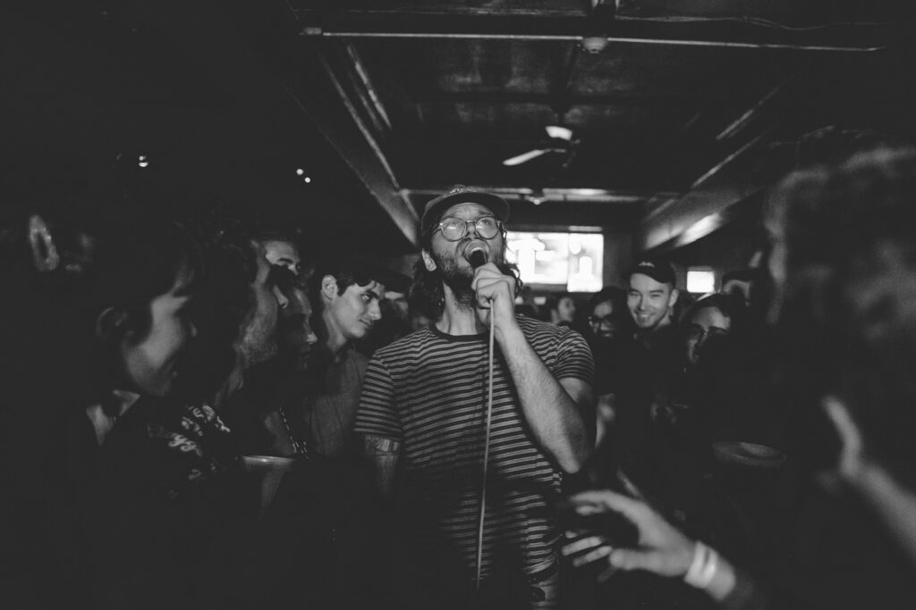 A singer wearing glasses and a striped shirt performs passionately with a microphone amid a crowd in a dimly lit venue. People around are smiling and enjoying the music. The image is in black and white.